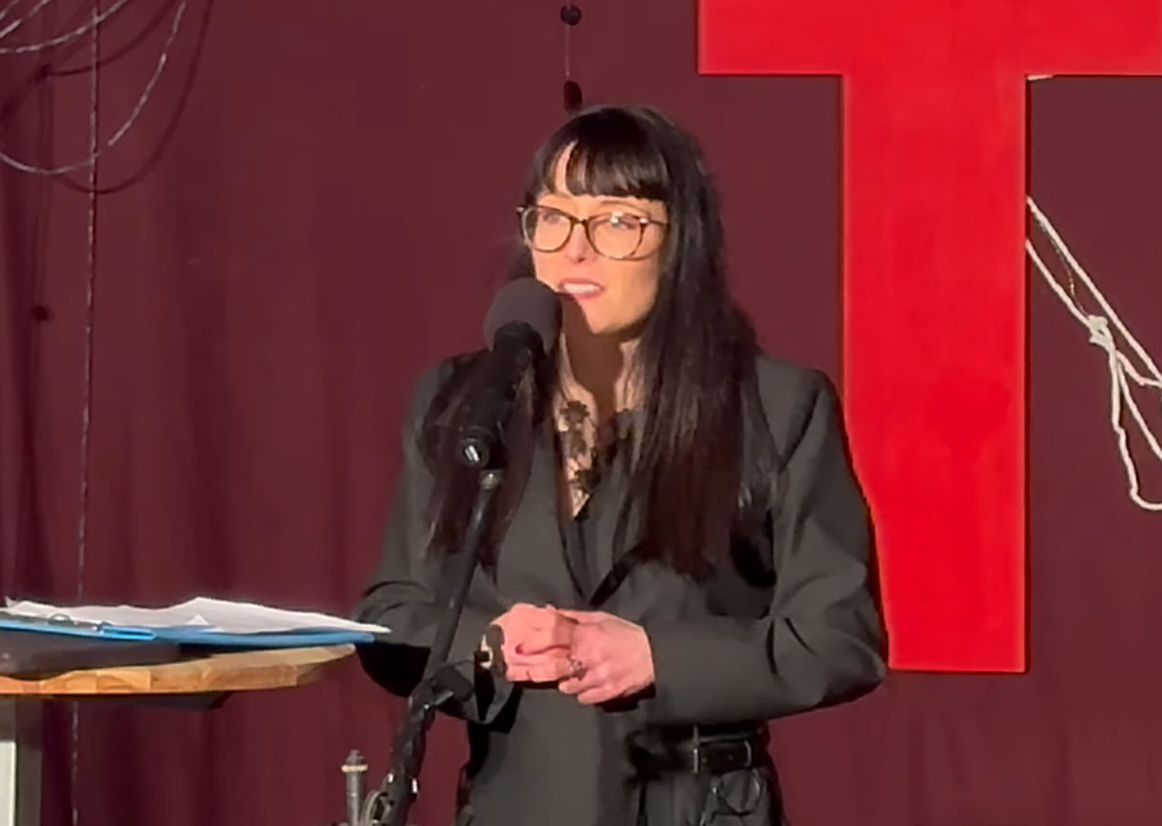 Lauren Muratore standing on stage with black dress and glasses in front of tedx logo.